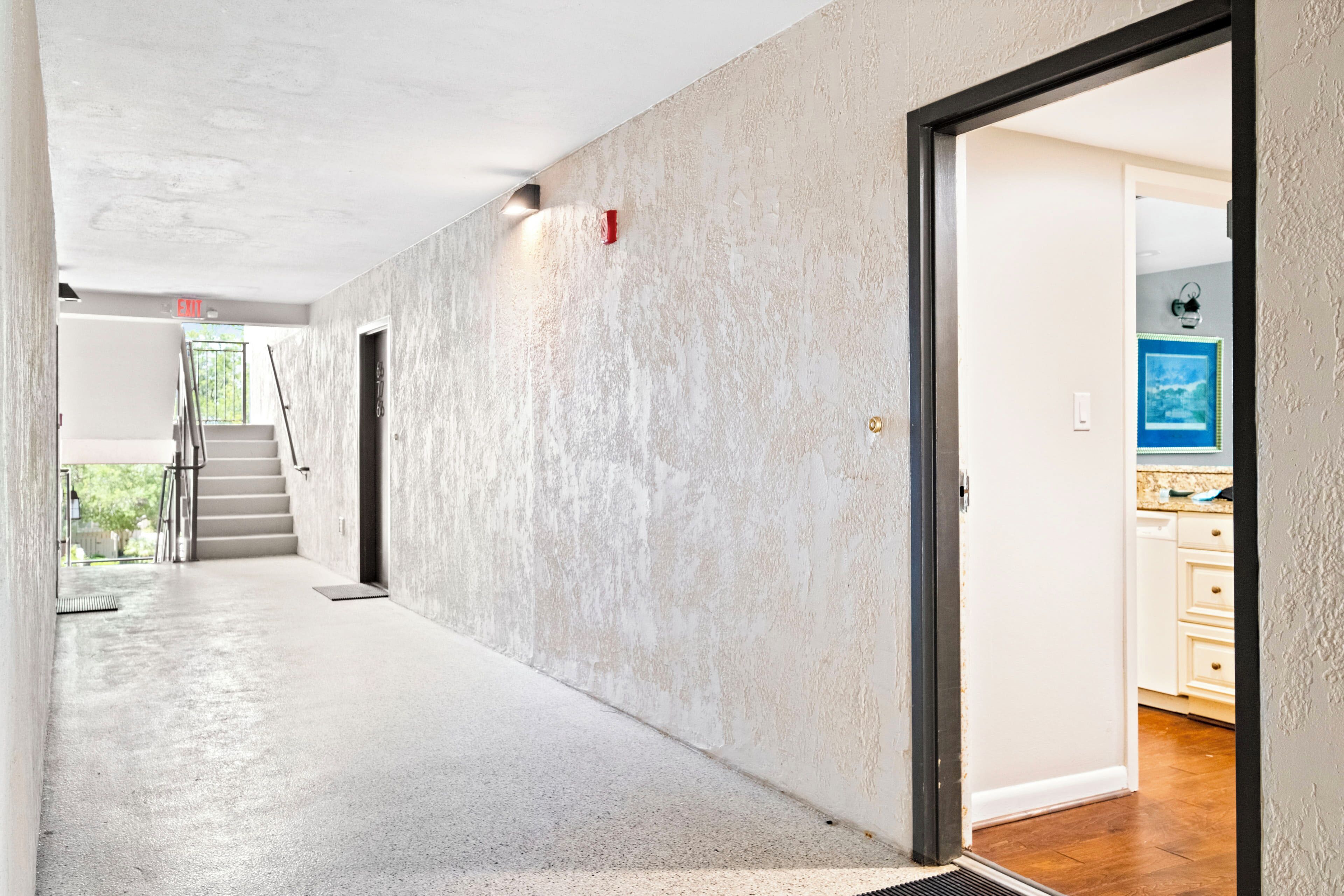 A hallway with a concrete floor and walls, leading to a kitchen area with wooden cabinets and a blue framed picture on the wall.