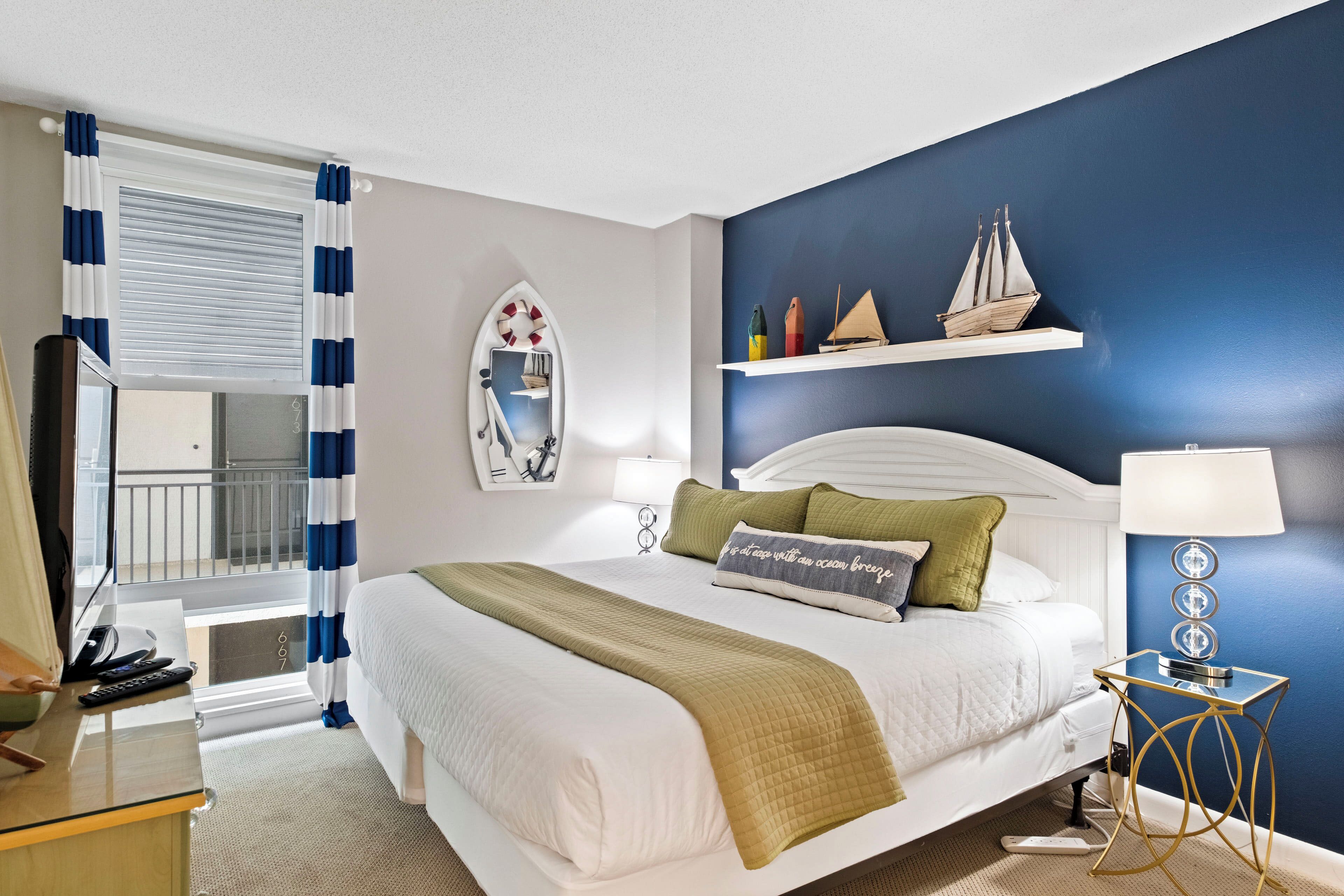 A hotel room with a bed, a desk, a chair, a mirror, a window with curtains, a nightstand, and a wall with a shelf holding decorative items.