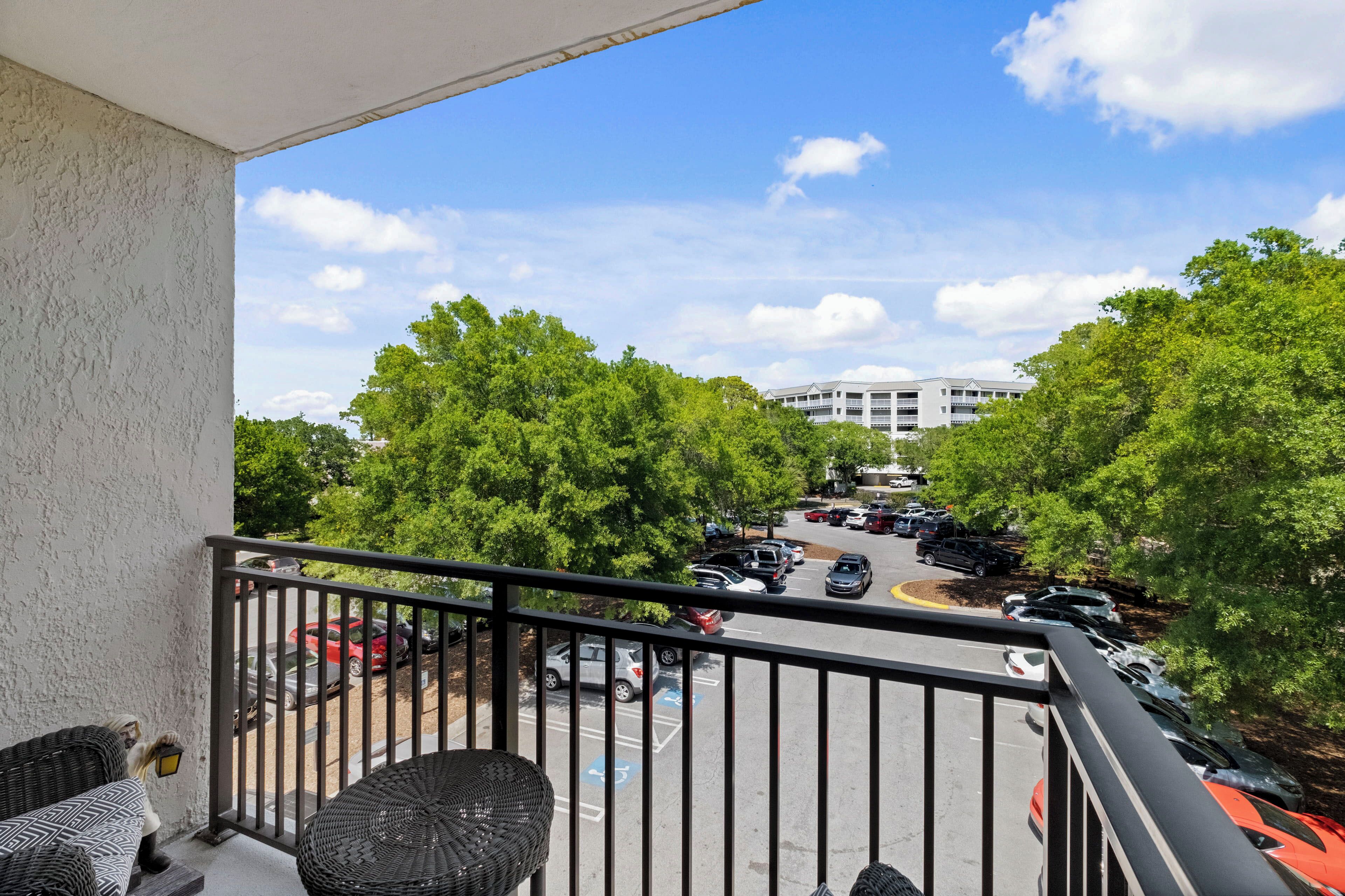 A balcony with wicker furniture, a view of a parking lot with cars, and a building in the background.
