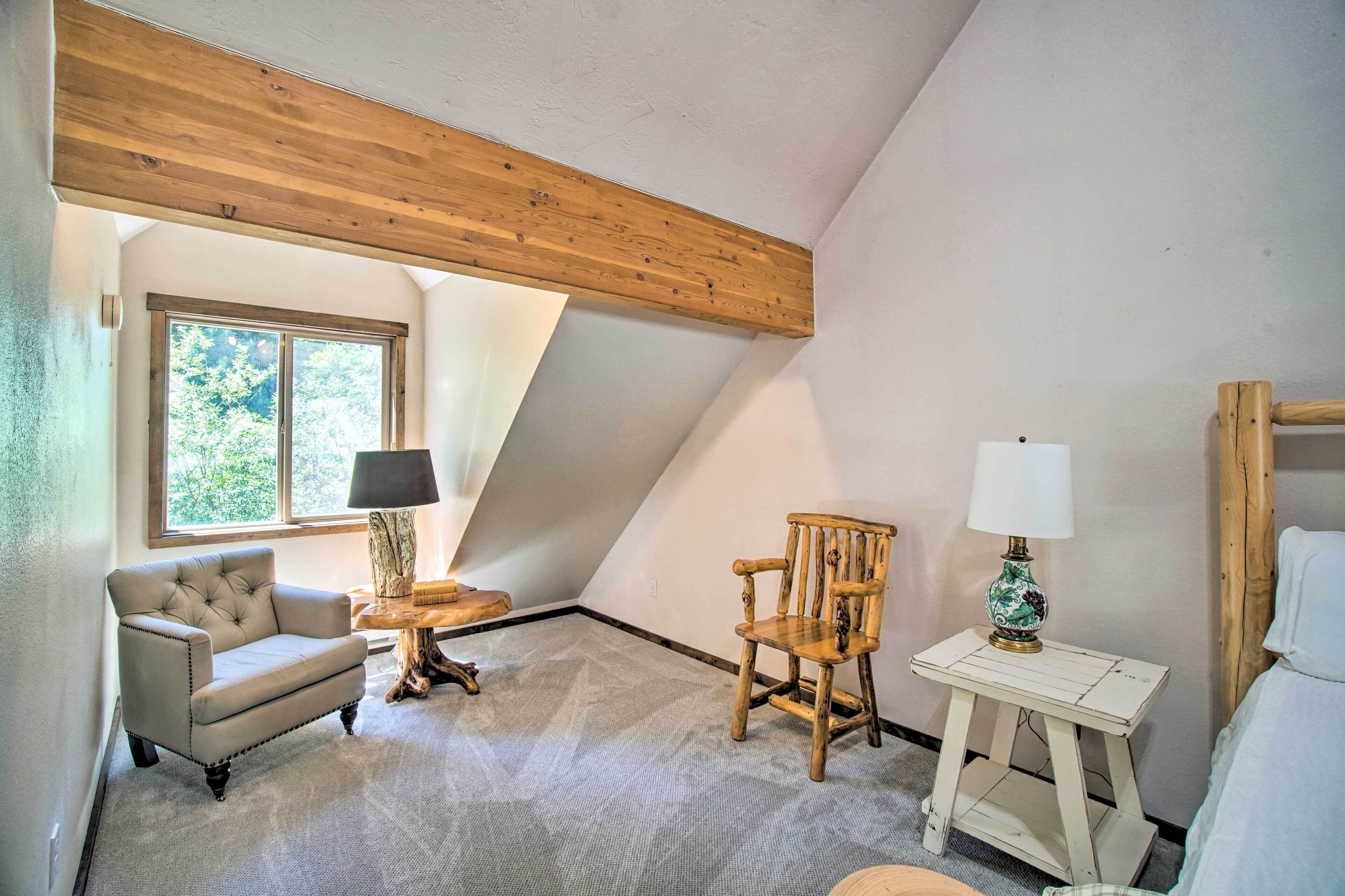 Ein gemütliches Zimmer mit hoher Decke, einem großen Fenster, einem gepolsterten Sessel, einem hölzernen Schaukelstuhl und einem kleinen Tisch mit Lampe.