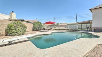 A swimming pool with a diving board, surrounded by a white fence, and a red umbrella in the background.