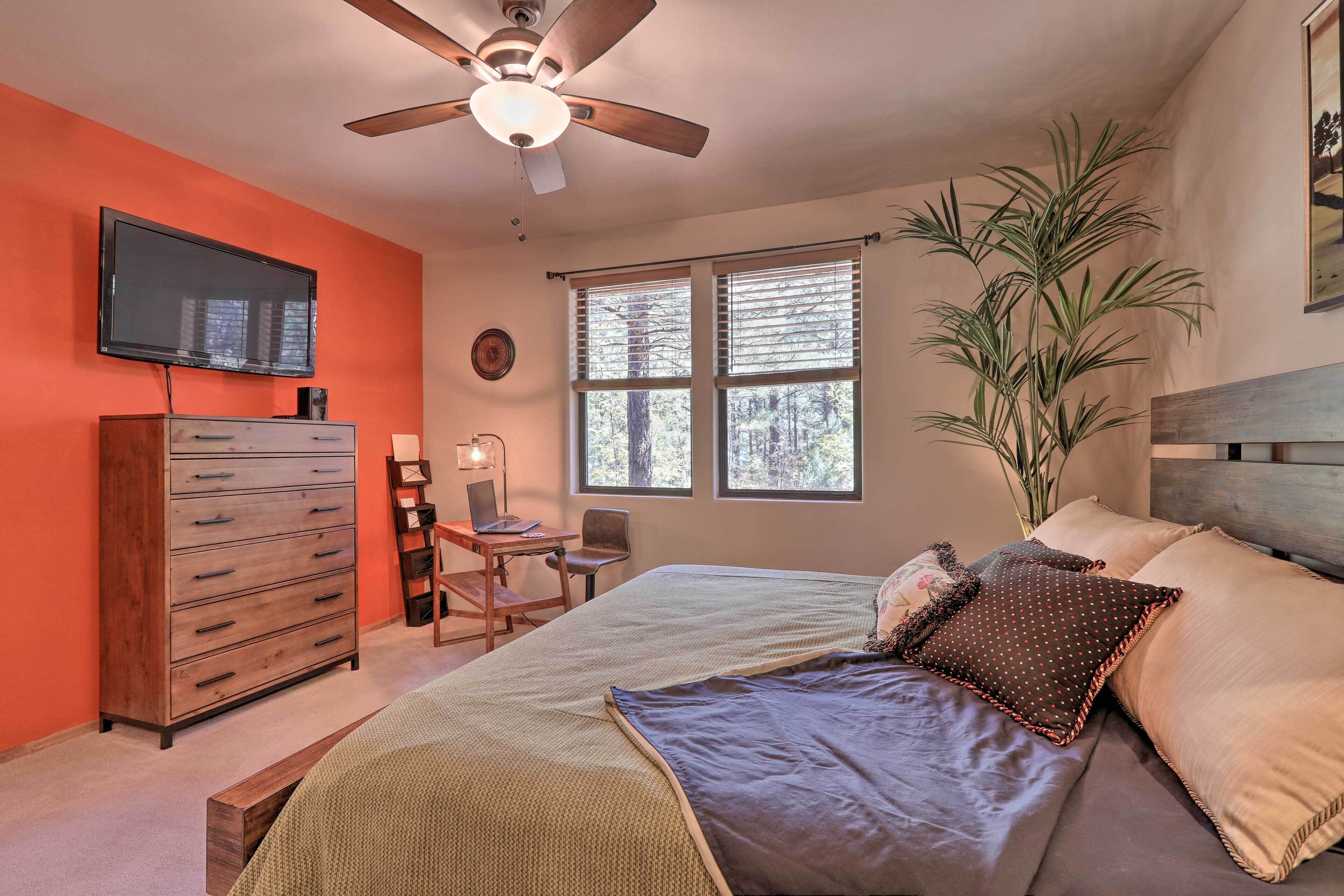 A bedroom with a bed, a dresser, a TV, a desk with a chair, a window, and a ceiling fan.