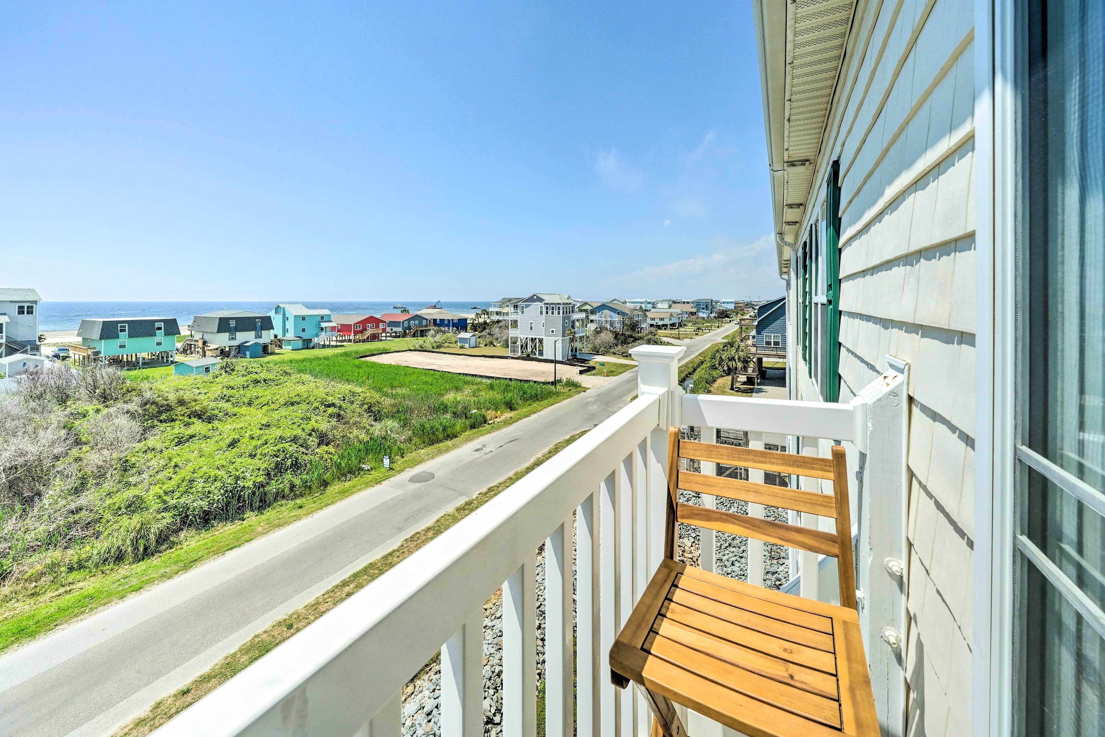 A balcony with a wooden chair, overlooking a road and beach houses, with a clear view of the ocean.