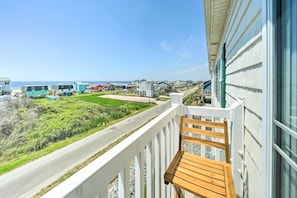 A balcony with a wooden chair, overlooking a road and beach houses, with a clear view of the ocean.