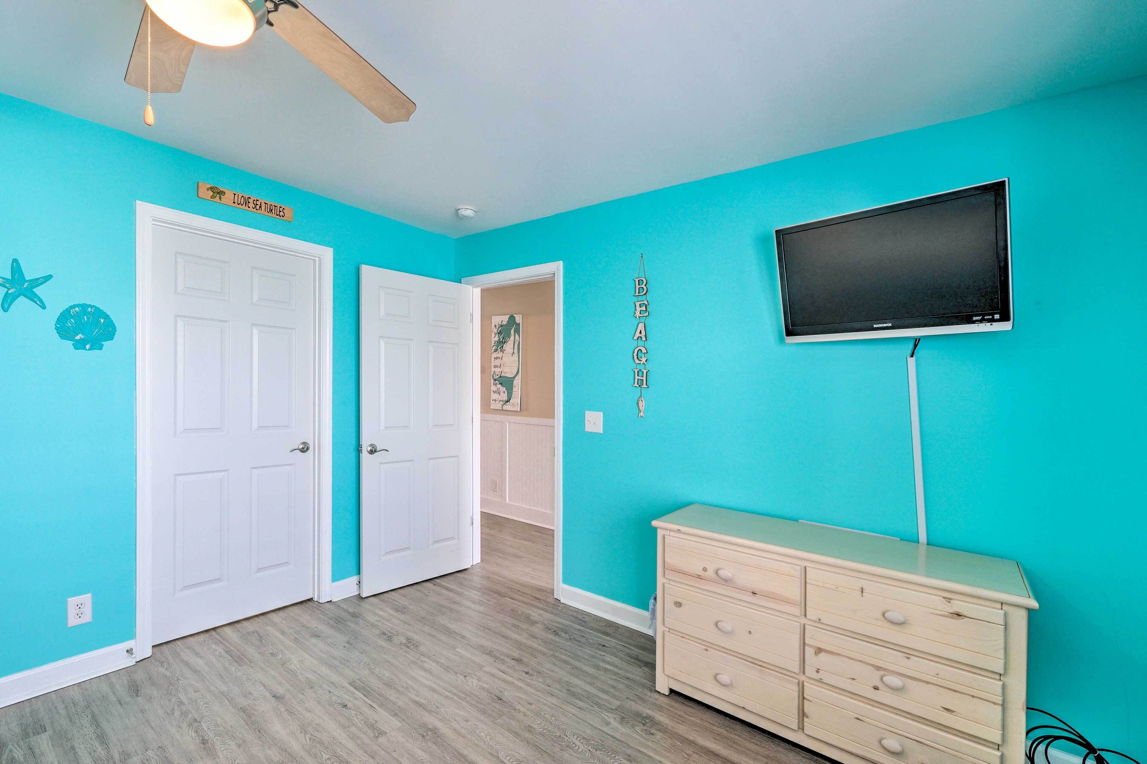 A room with turquoise walls, a ceiling fan, a TV mounted on the wall, a wooden dresser, and a door with a sign.