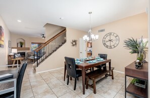 A dining area with a wooden table, chairs, and a chandelier. There is a staircase leading to another room.
