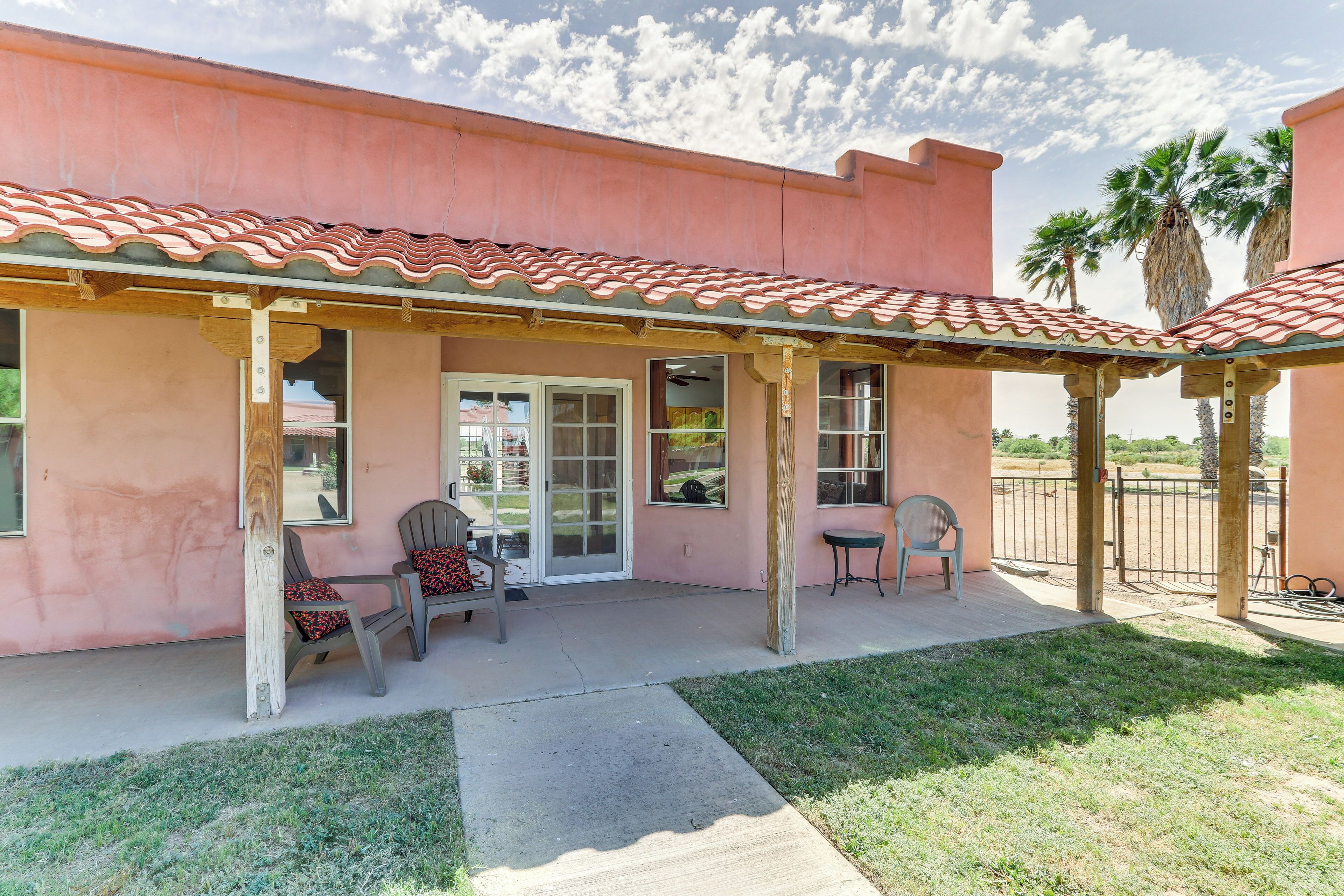 A covered patio with chairs and a table, a building with a red-tiled roof, and palm trees in the background.