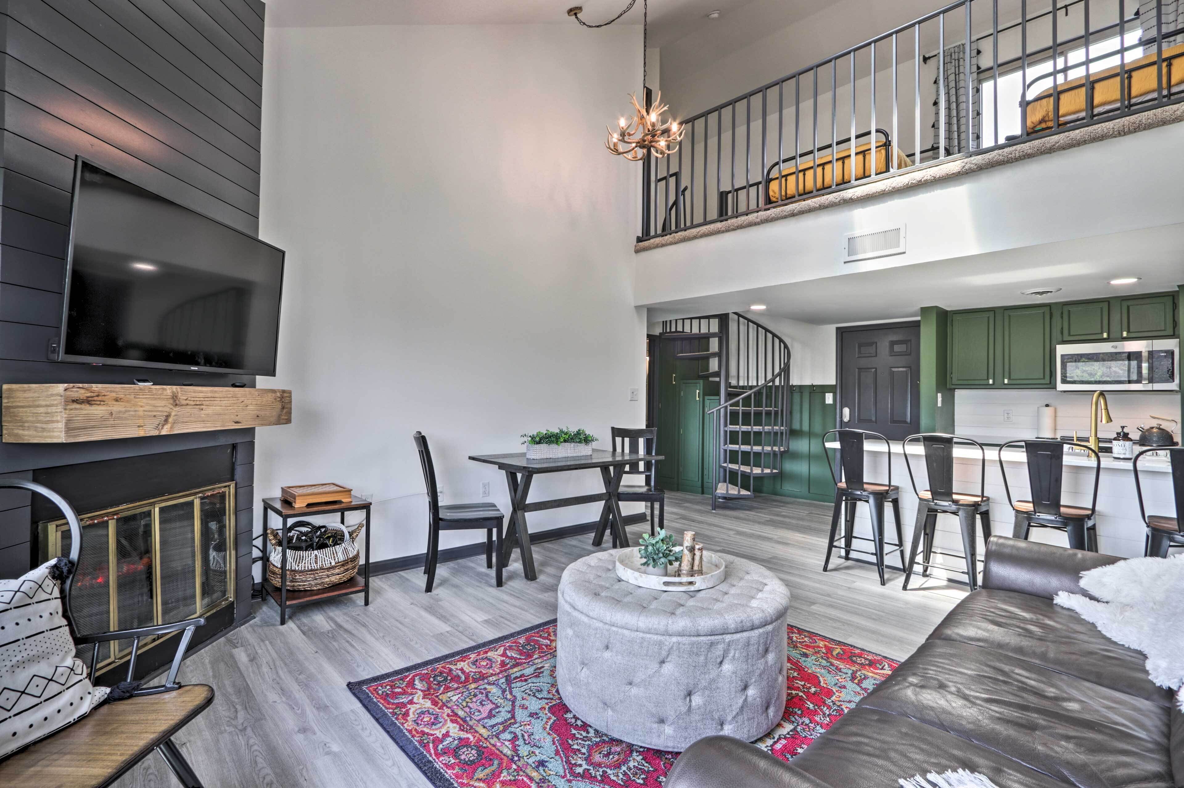 A spacious living area with a fireplace, a television, and a dining set. A staircase leads to an upper level with a balcony.