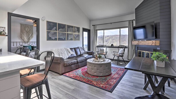 A modern living room with a sofa, ottoman, dining table, and chairs. There is a fireplace, a TV mounted on the wall, and a large window with a view.