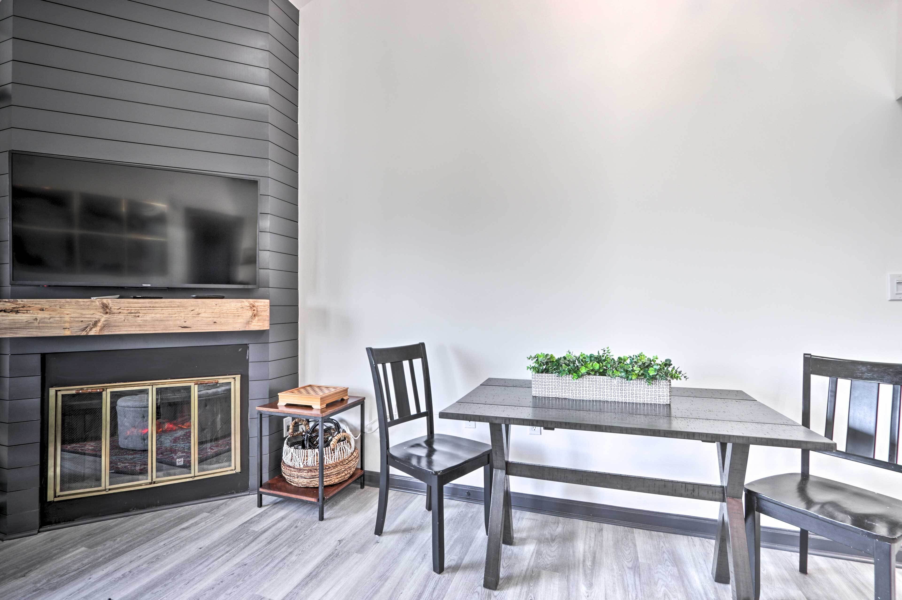 Uma sala de jantar com uma mesa de madeira escura, duas cadeiras, uma lareira com uma TV acima e um cesto de plantas sobre a mesa.