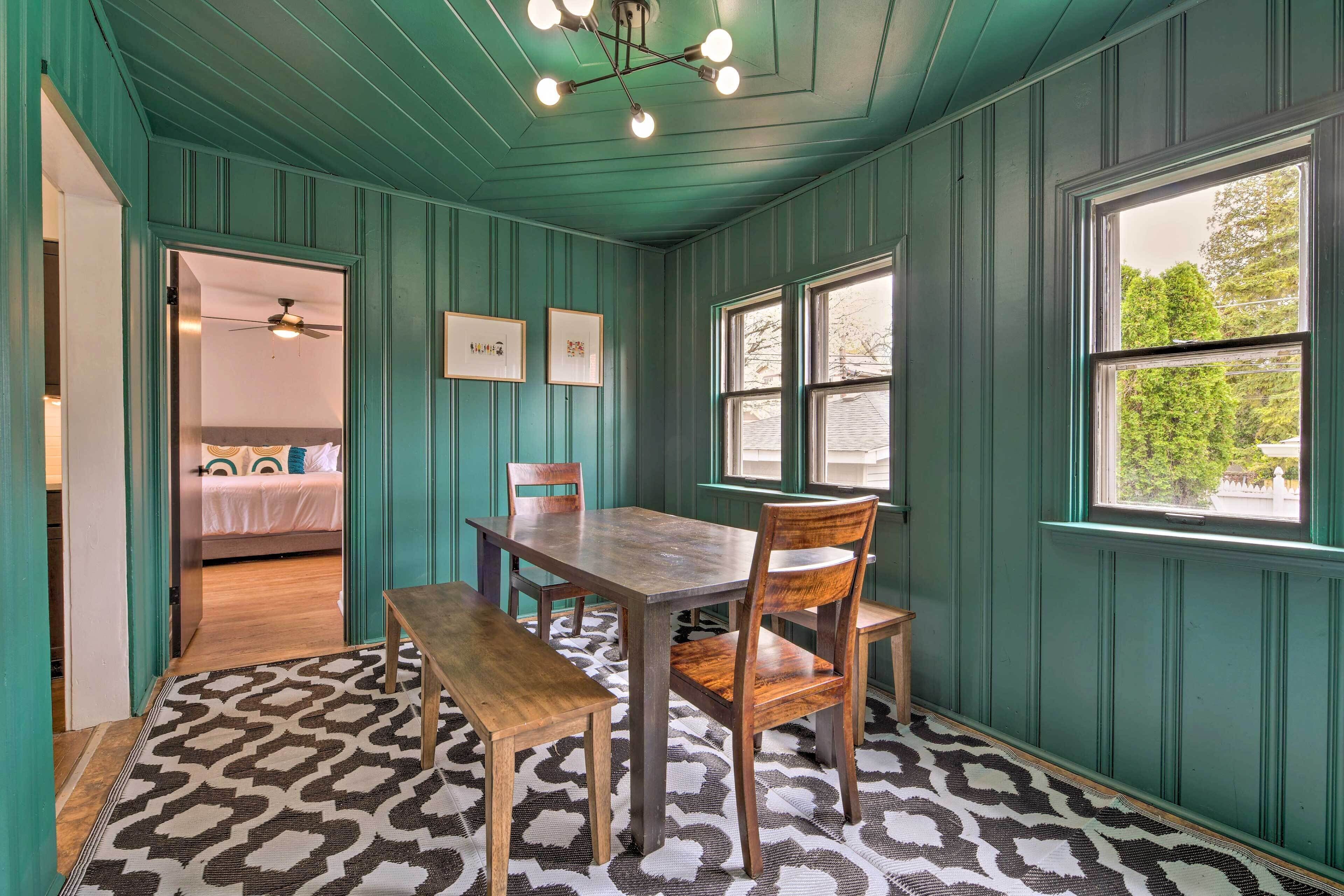 A room with teal walls, a wooden dining table with chairs, a patterned rug, and a window with a view of trees.