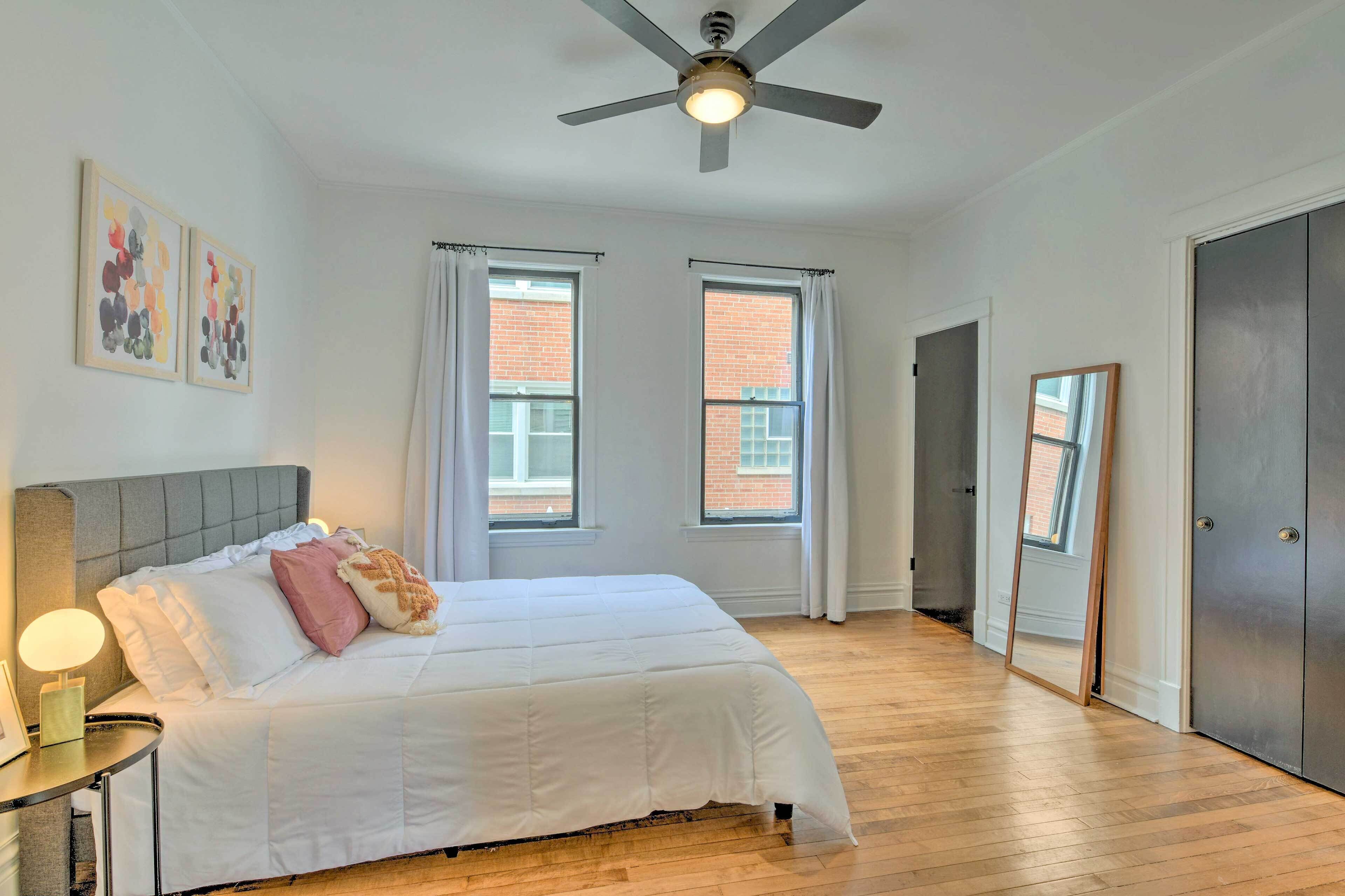 Quarto com cama, ventilador de teto, duas janelas com cortinas, porta e espelho.