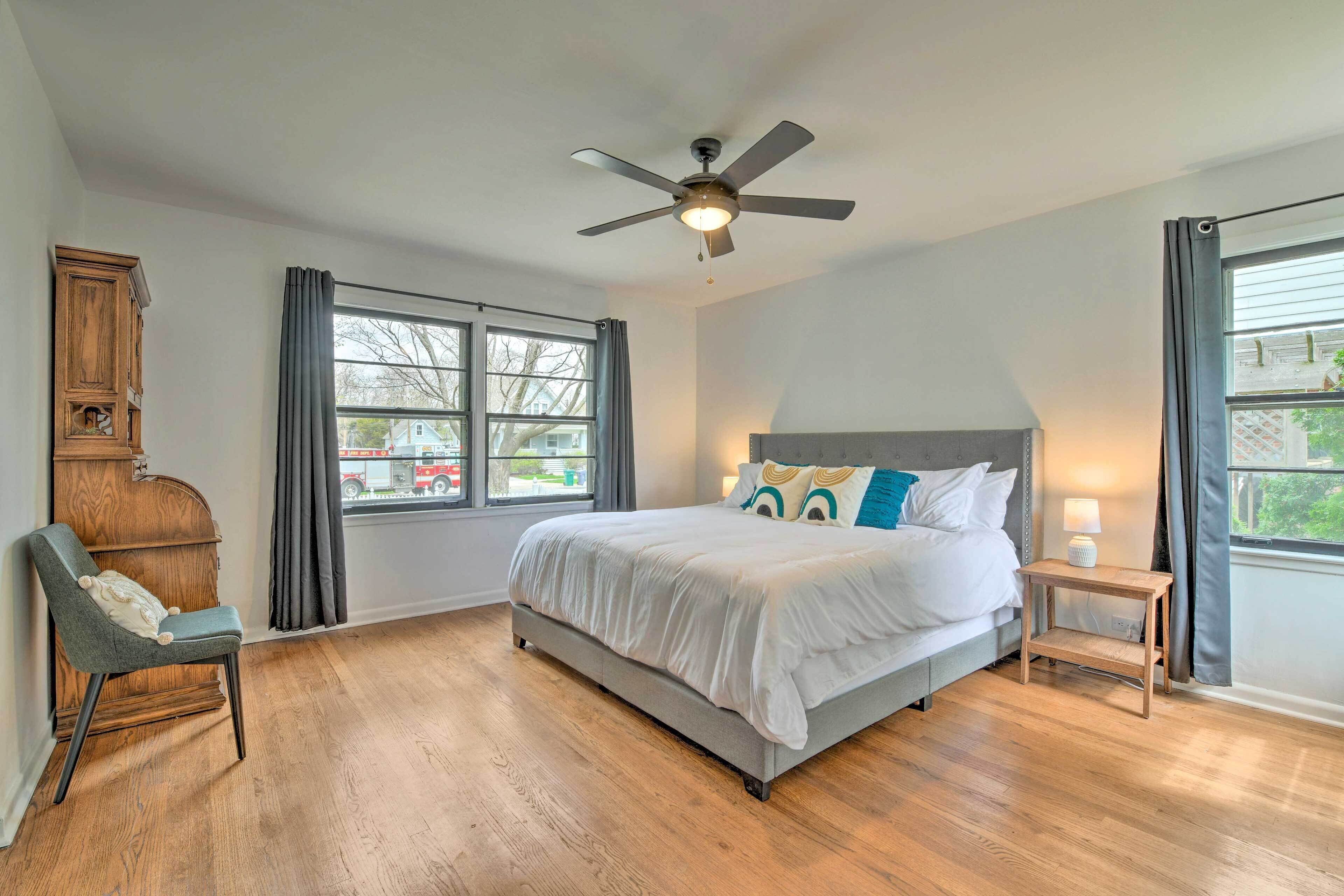 A bedroom with a bed, a chair, a nightstand, a ceiling fan, and large windows.