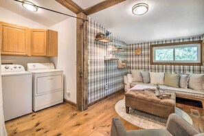 A laundry area with a washing machine and dryer, a plaid-patterned wall, a window with a view of trees, and a cozy seating area with a sofa and ottoman.