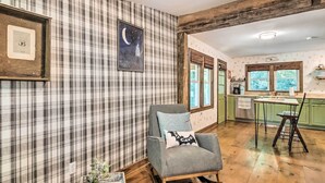 A cozy room with plaid wallpaper, a rocking chair, a wooden table, and a kitchen area with a window. - Whimsical Bat Cave Cottage w/ Fire Pit! (Hendersonville)