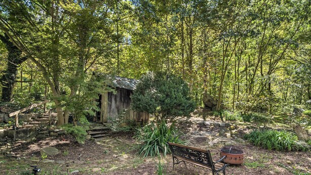 A rustic wooden cabin surrounded by dense forest, a stone bench, and a clay pot. - Whimsical Bat Cave Cottage w/ Fire Pit! (Hendersonville)