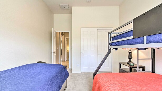 Quarto com duas camas, uma com colcha azul e a outra com colcha vermelha, uma mesinha de cabeceira com abajur, e uma porta que dá para outro cômodo.