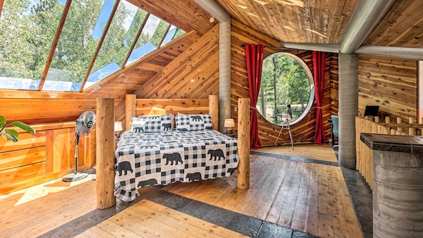 A cozy wooden cabin with a bed, a fan, and a circular window overlooking the forest. - Angel Fire Cabin w/ Deck ~ 2 Mi to Ski & Hiking! (Angel Fire)