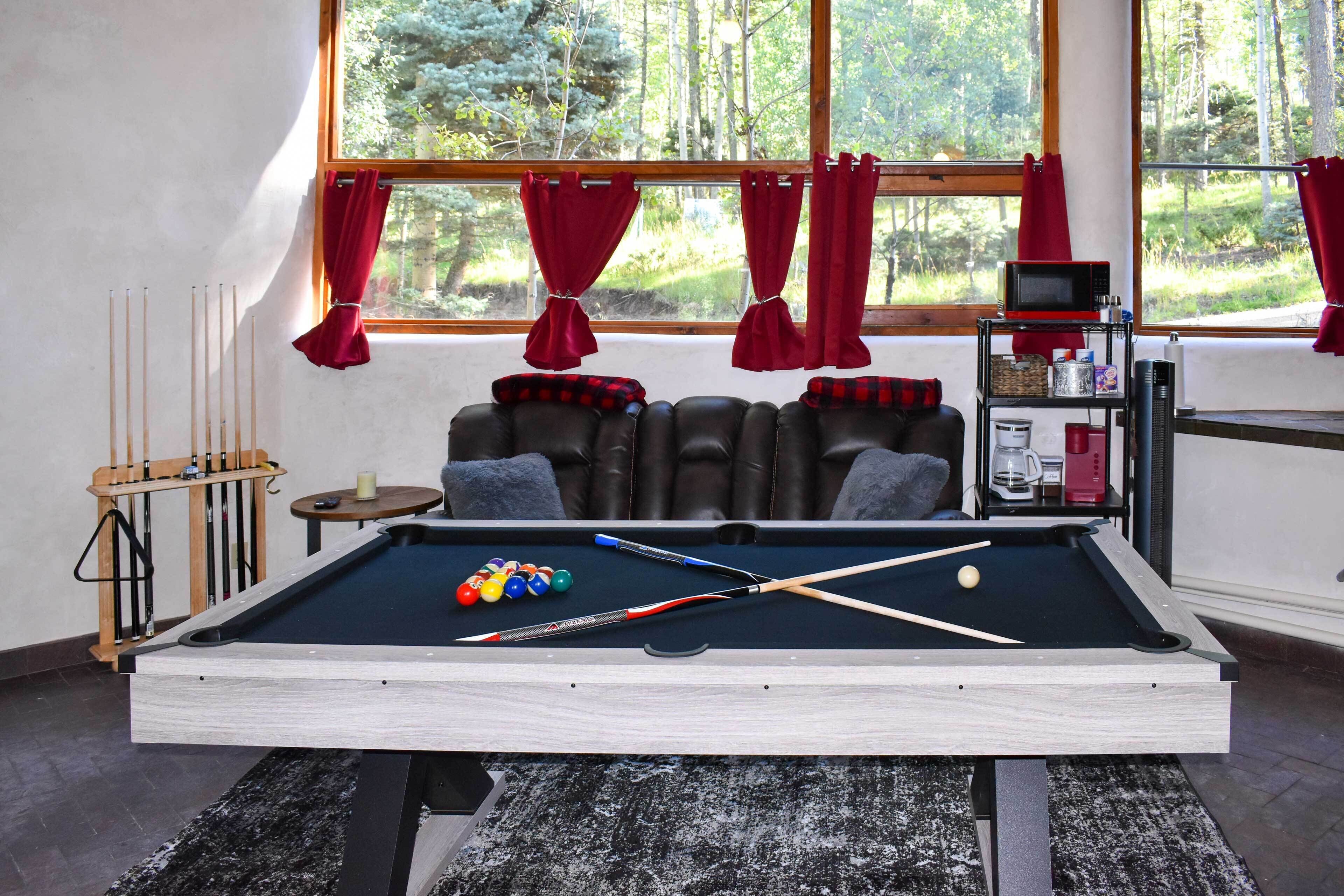 A pool table in a room with a view of a forest through large windows, a bookshelf with various items, and a billiards rack.