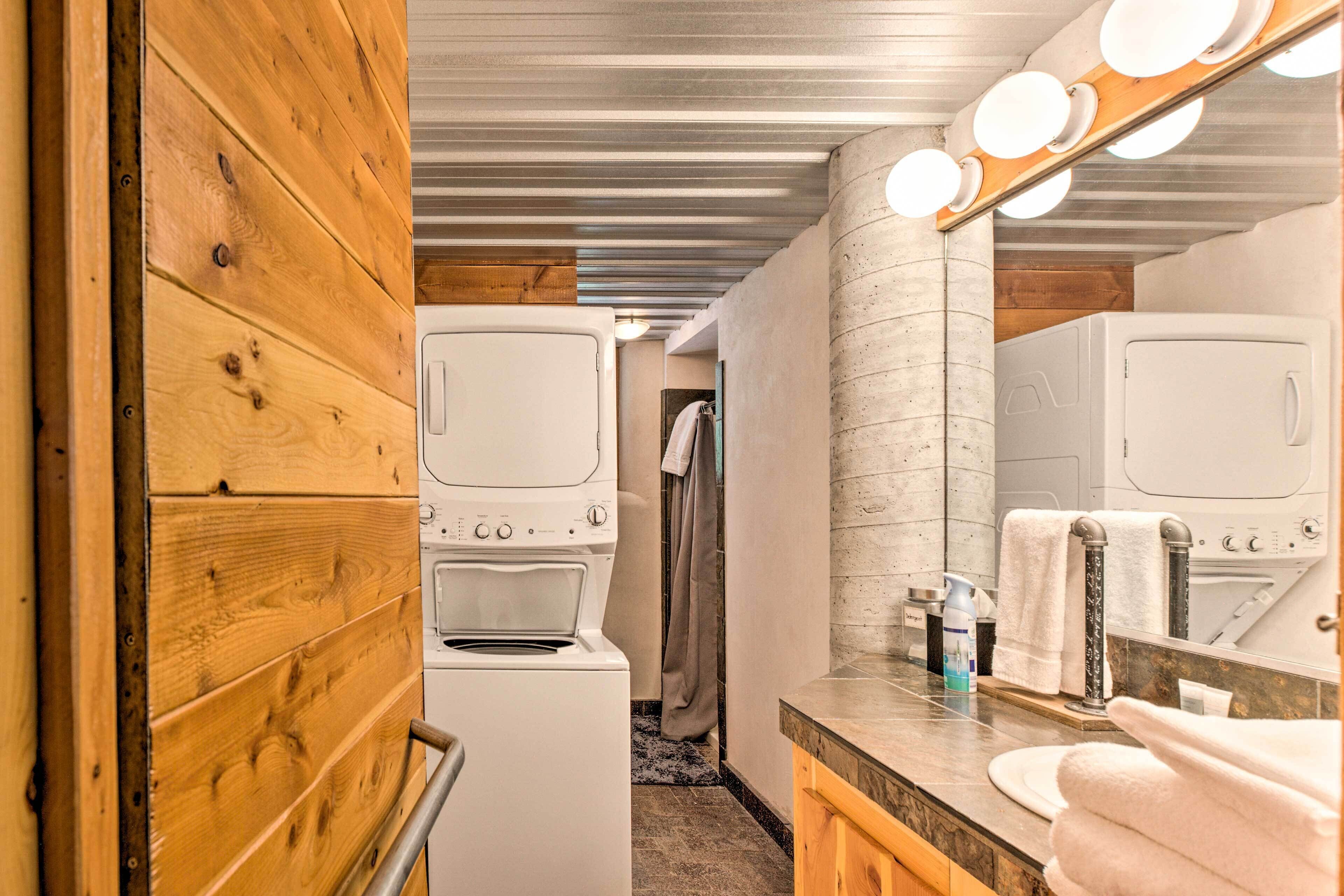 A compact bathroom with a wooden wall, a white washing machine, a mirror with lights, and a sink with a towel.