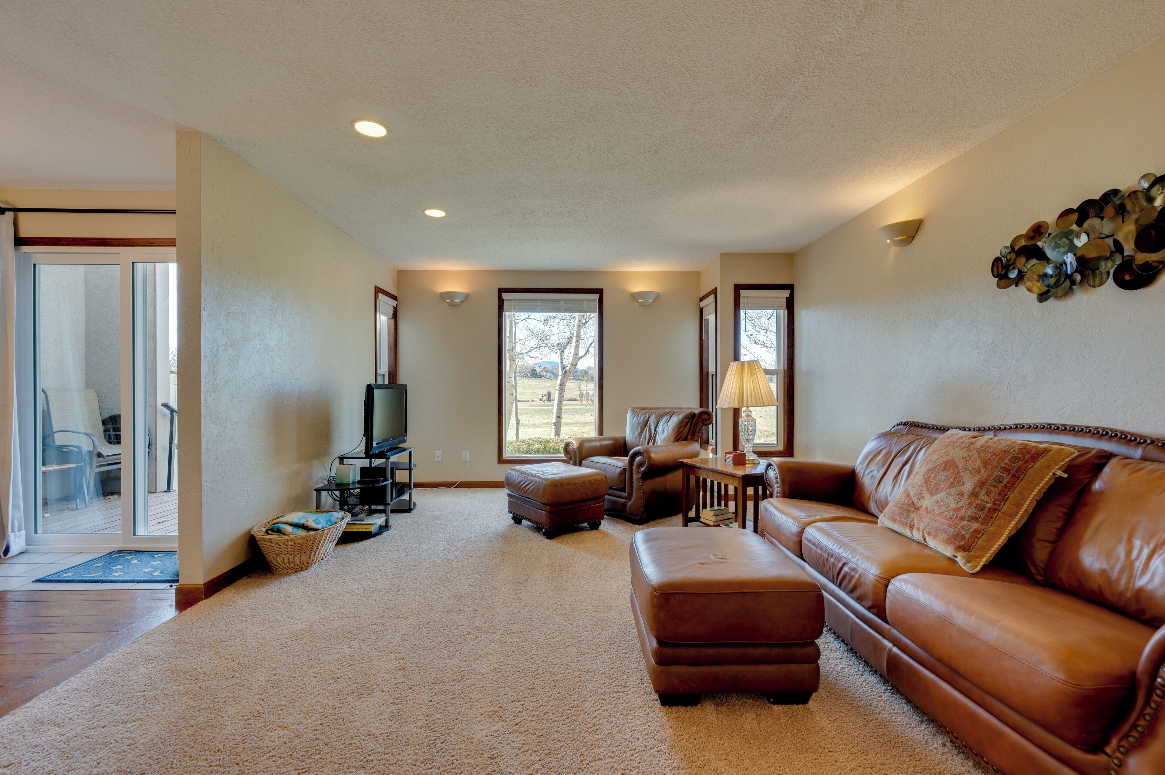 A spacious living room with a leather sofa, ottoman, and a fireplace. There is a large window providing natural light and a view of the outdoors.