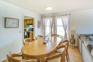 A dining area with a wooden table and chairs, a kitchen in the background, and a living room with a sofa and a lamp.