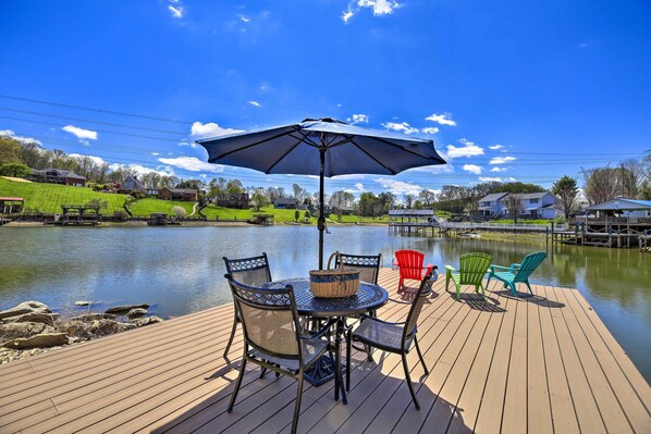 A wooden deck with a table and chairs, an umbrella, and a view of a lake and houses. - Lake House Haven: Fire Pit, Boat Dock + More! (Johnson City)