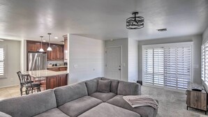 A modern living room with a sectional sofa, a kitchen area with wooden cabinets, a stainless steel refrigerator, pendant lights, and a large window with blinds. - Parker Home on the Colorado River w/ Views! (Parker)