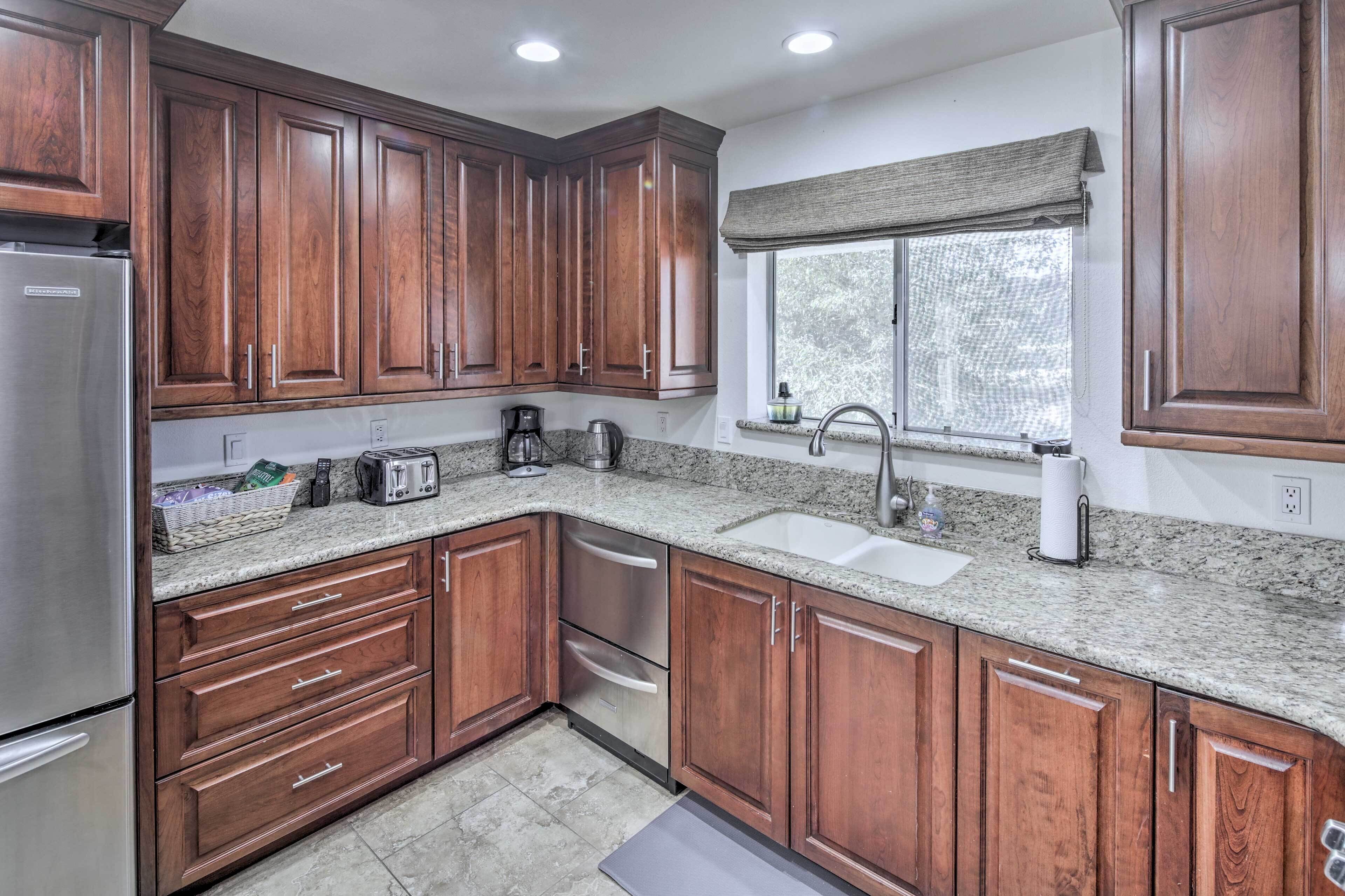 A kitchen with wooden cabinets, granite countertops, a stainless steel refrigerator, a coffee maker, and a window with a view of trees.