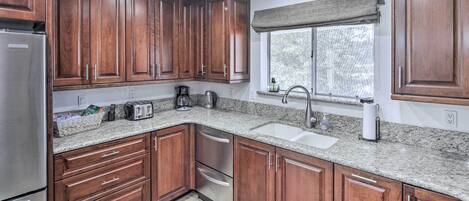 A kitchen with wooden cabinets, granite countertops, a stainless steel refrigerator, a coffee maker, and a window with a view of trees.