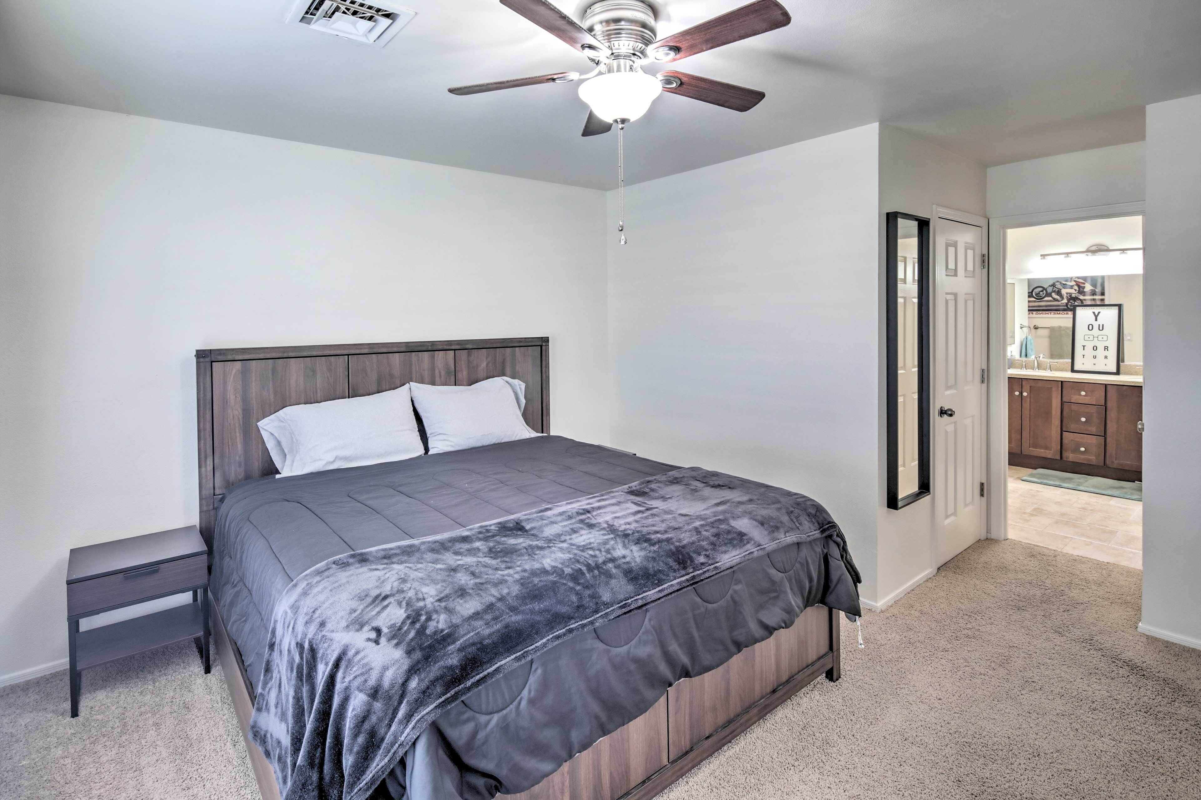 Une chambre à coucher comprenant un lit, une table de chevet, un ventilateur de plafond et une porte donnant sur une salle de bain.