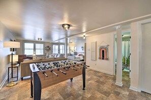 A spacious living area with a foosball table, a sofa, and a coffee table. There are multiple windows providing natural light, and a hallway leading to other rooms.