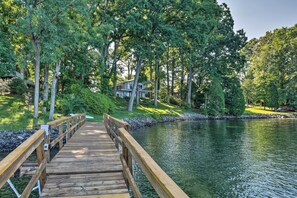 Un pont en bois qui mène à une maison entourée d’arbres et bordée d’un lac.