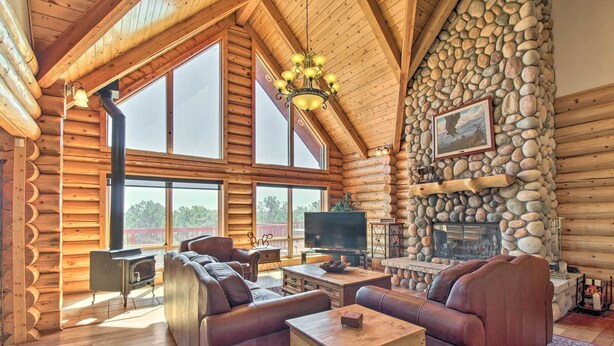 A log cabin interior with a stone fireplace, leather sofas, a television, and a large window. - Hot Tub, Game Room & Theater: Luxe Show Low Home (Show Low)