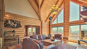 A log cabin interior with a large leather sofa, wooden walls, and a large window offering a view. - Hot Tub, Game Room & Theater: Luxe Show Low Home (Show Low)