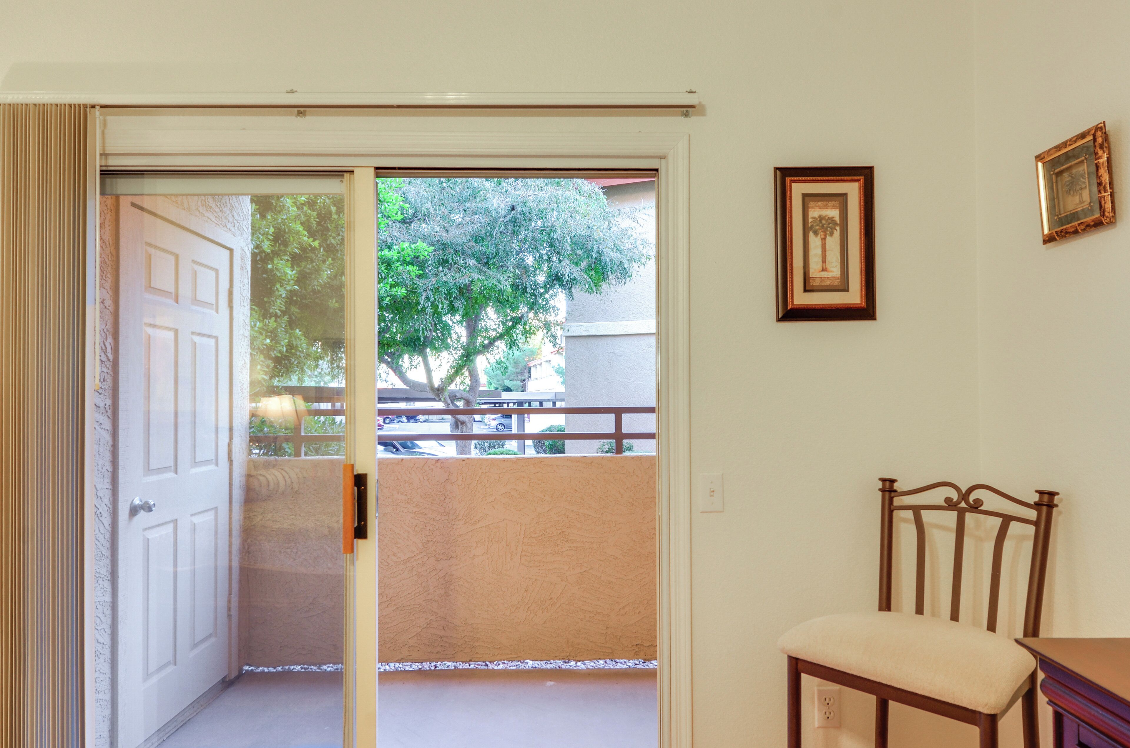 A room with a sliding glass door leading to a balcony, a wooden chair, and two framed pictures on the wall.