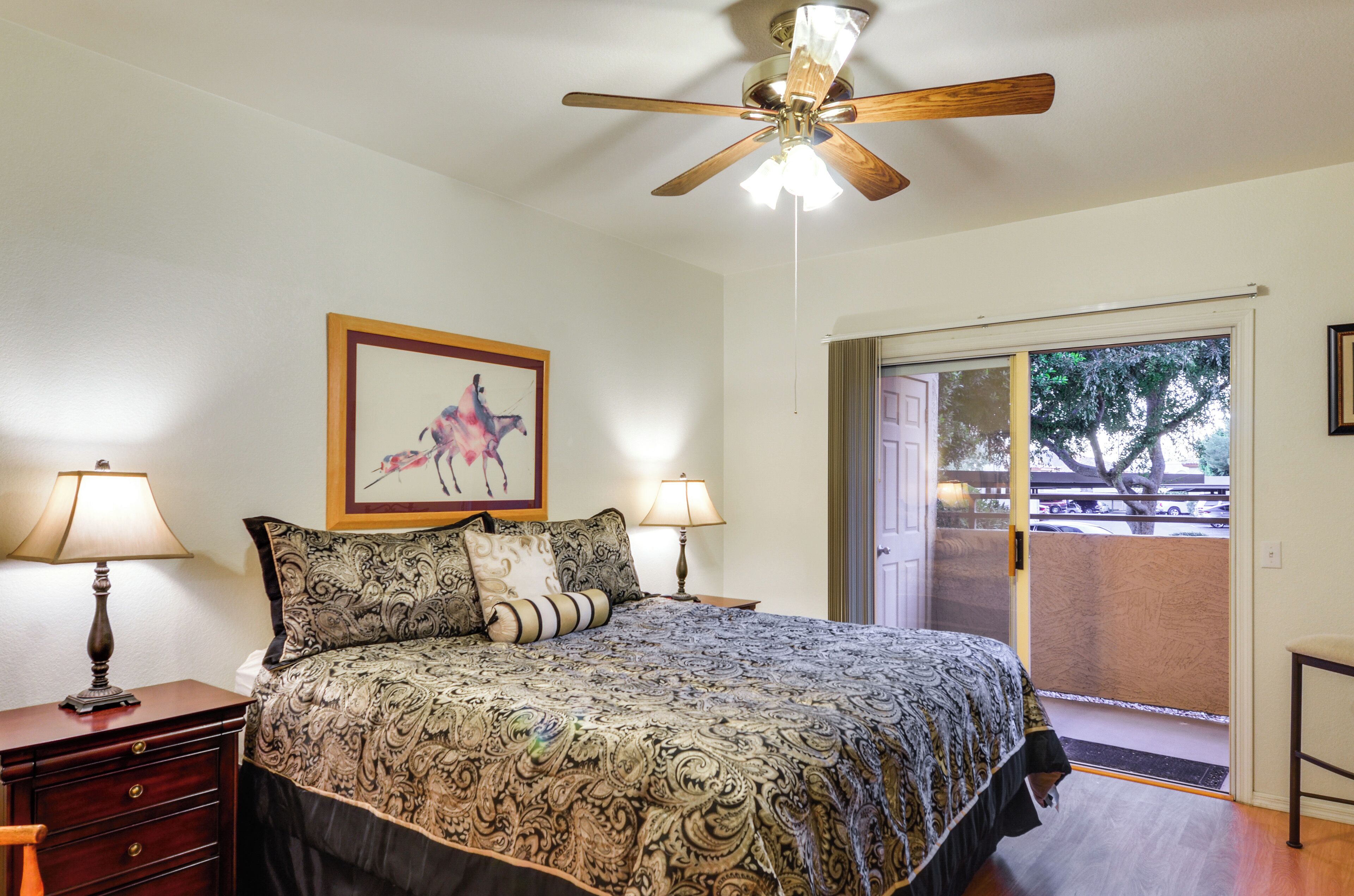 A bedroom with a bed, two bedside lamps, a ceiling fan, a framed picture on the wall, and a view of the outside through a sliding door.