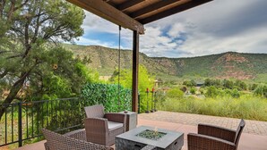 A covered patio with wicker furniture, a fire pit, and a view of green hills and a building in the distance. - Sedona Retreat w/ Patio, Bar & Swim Spa! (Sedona)