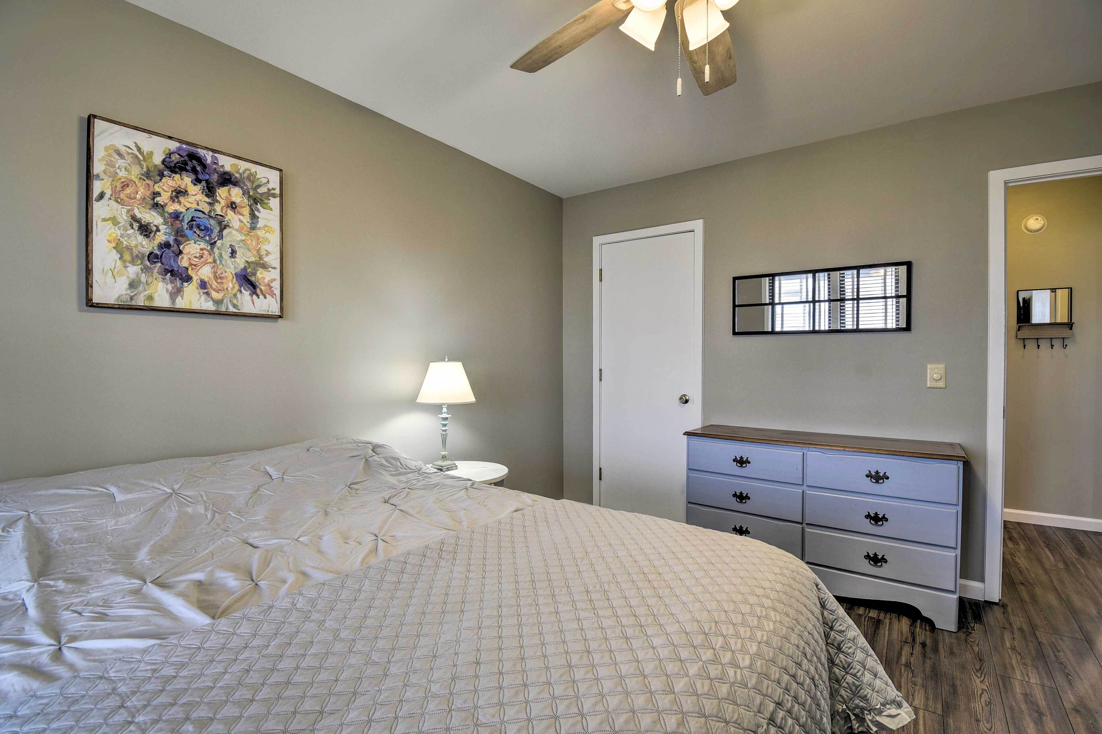 A bedroom with a bed, a dresser, a lamp, a ceiling fan, and a painting on the wall.