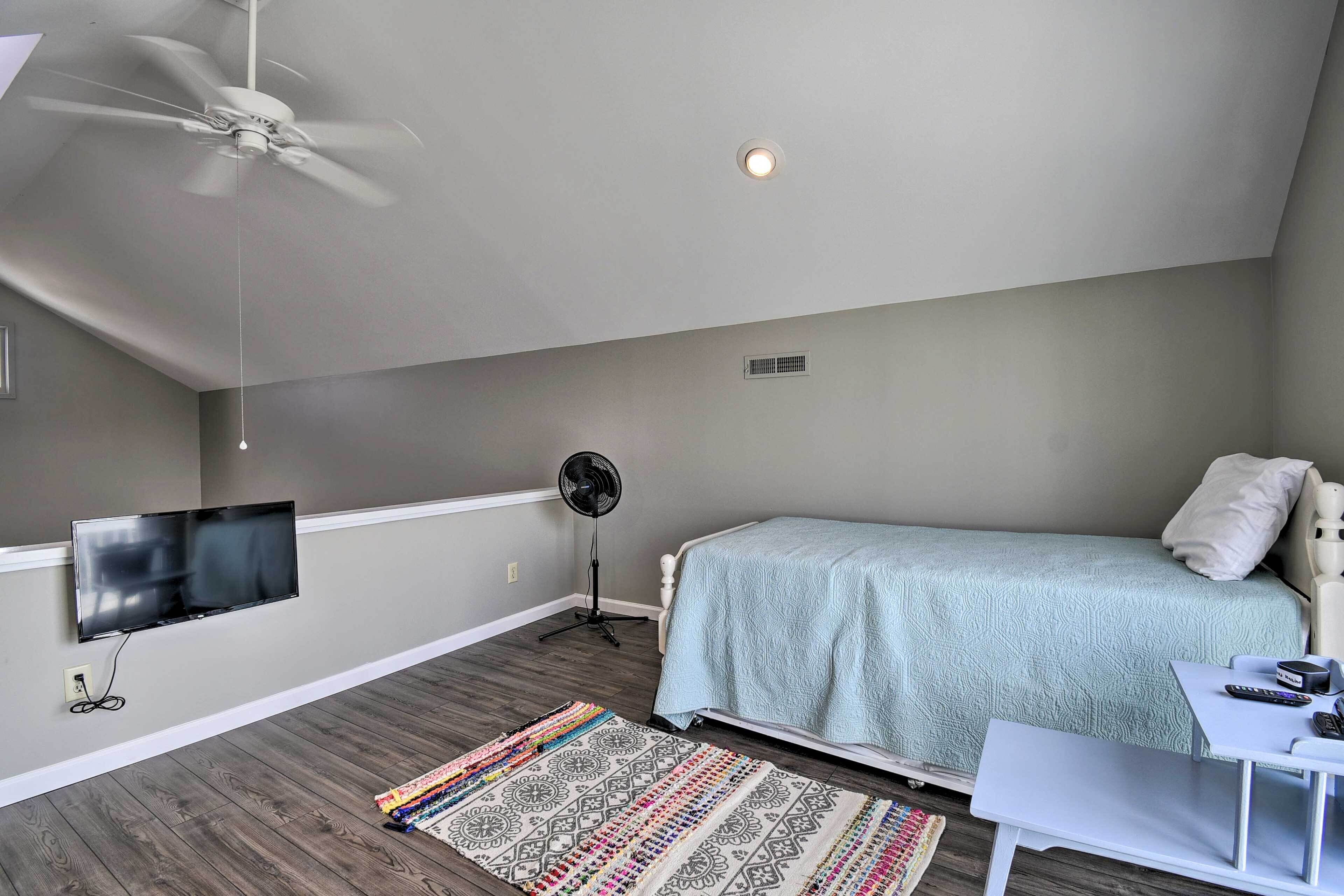 A bedroom with a bed, a TV mounted on the wall, a ceiling fan, a standing fan, and a small table with a remote control.