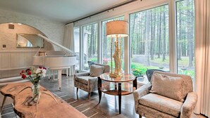 Un salón amplio con un piano de cola, un ventanal con vistas a un bosque y una lámpara de pie alta.