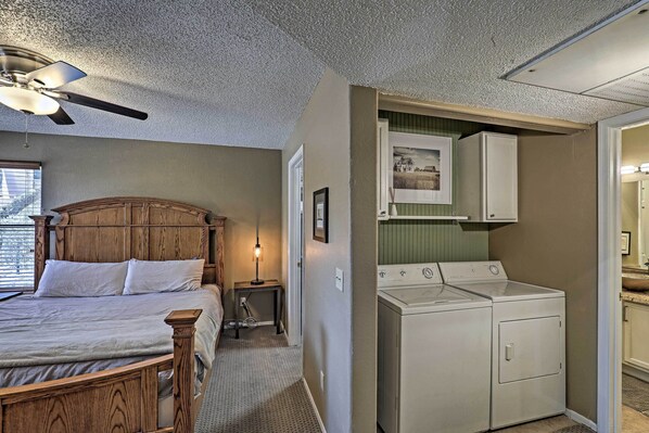 A bedroom with a wooden bed, a ceiling fan, a washing machine, and a small table with a lamp.