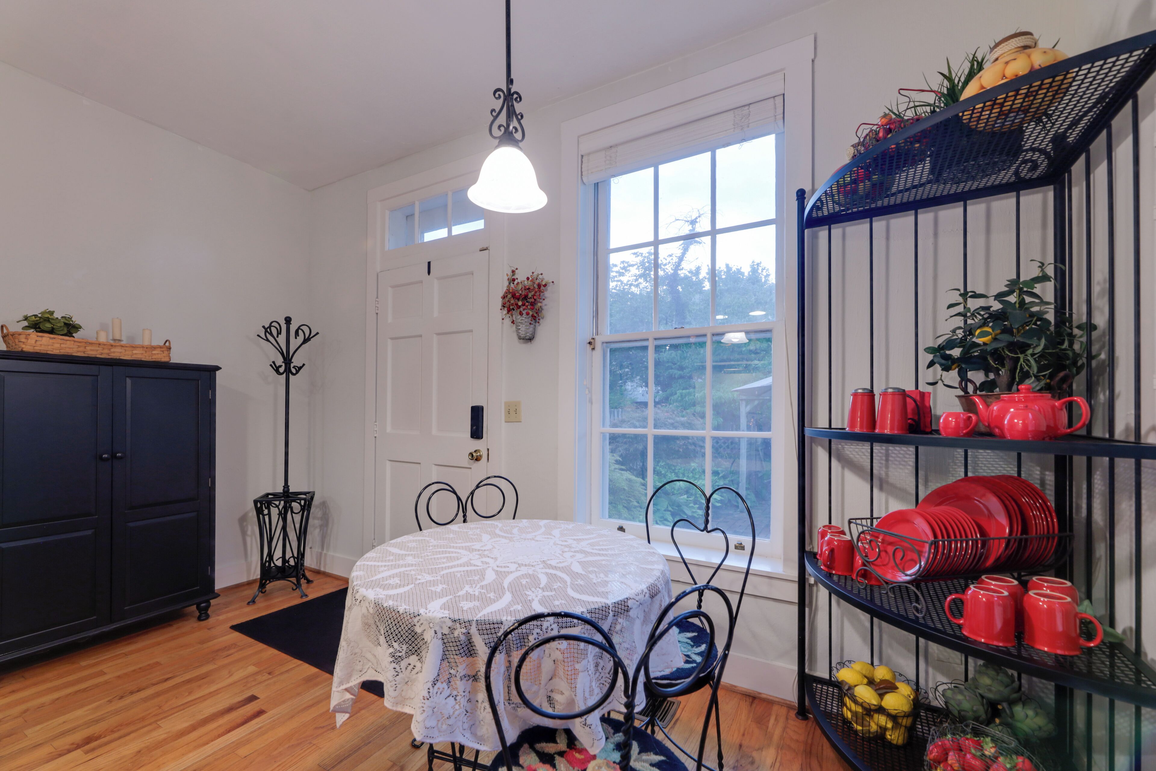 A dining area with a round table, chairs, and a chandelier. There is a black metal shelf with red dishes and a basket of fruit.