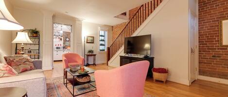 A living room with a sofa, armchair, coffee table, and TV. There's a staircase with a brick wall and a door leading to another room.