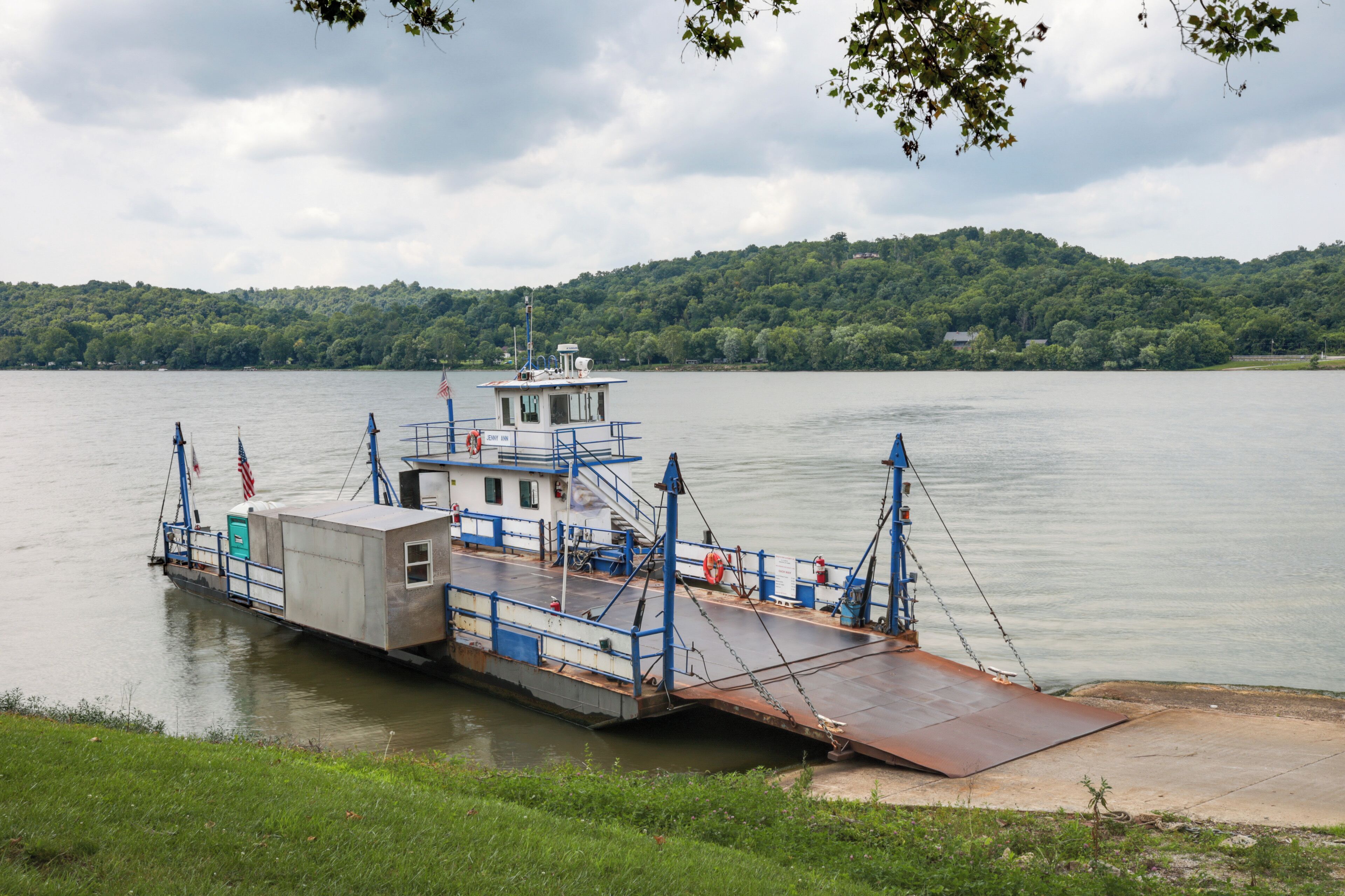Eine Fähre liegt an einem Fluss vor einer bewaldeten Hügelkette.