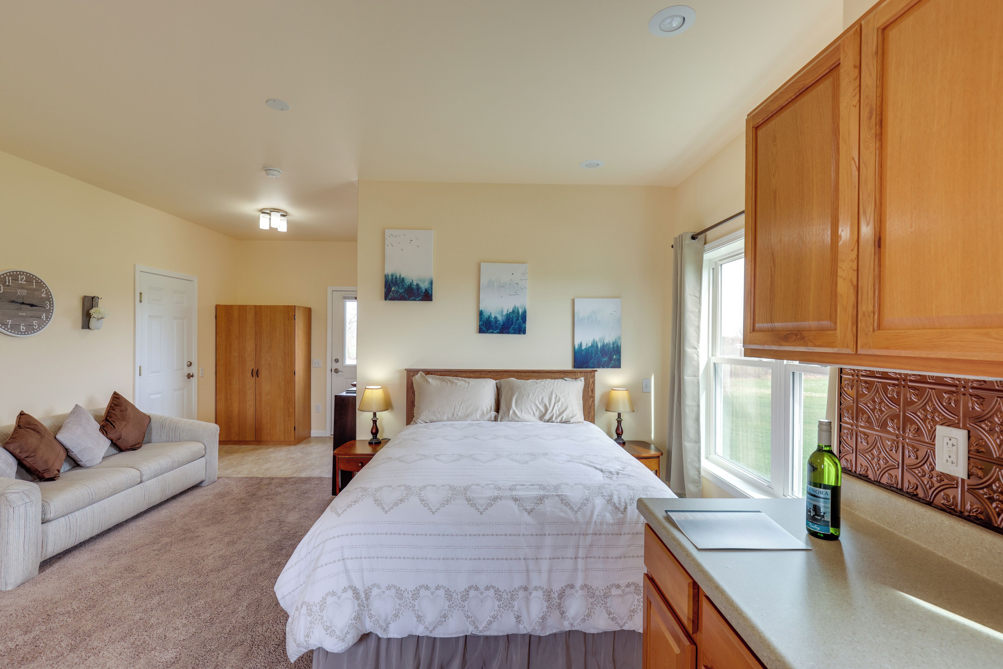 A bedroom with a large bed, a bedside table with lamps, a dresser, a window with curtains, and a clock on the wall.