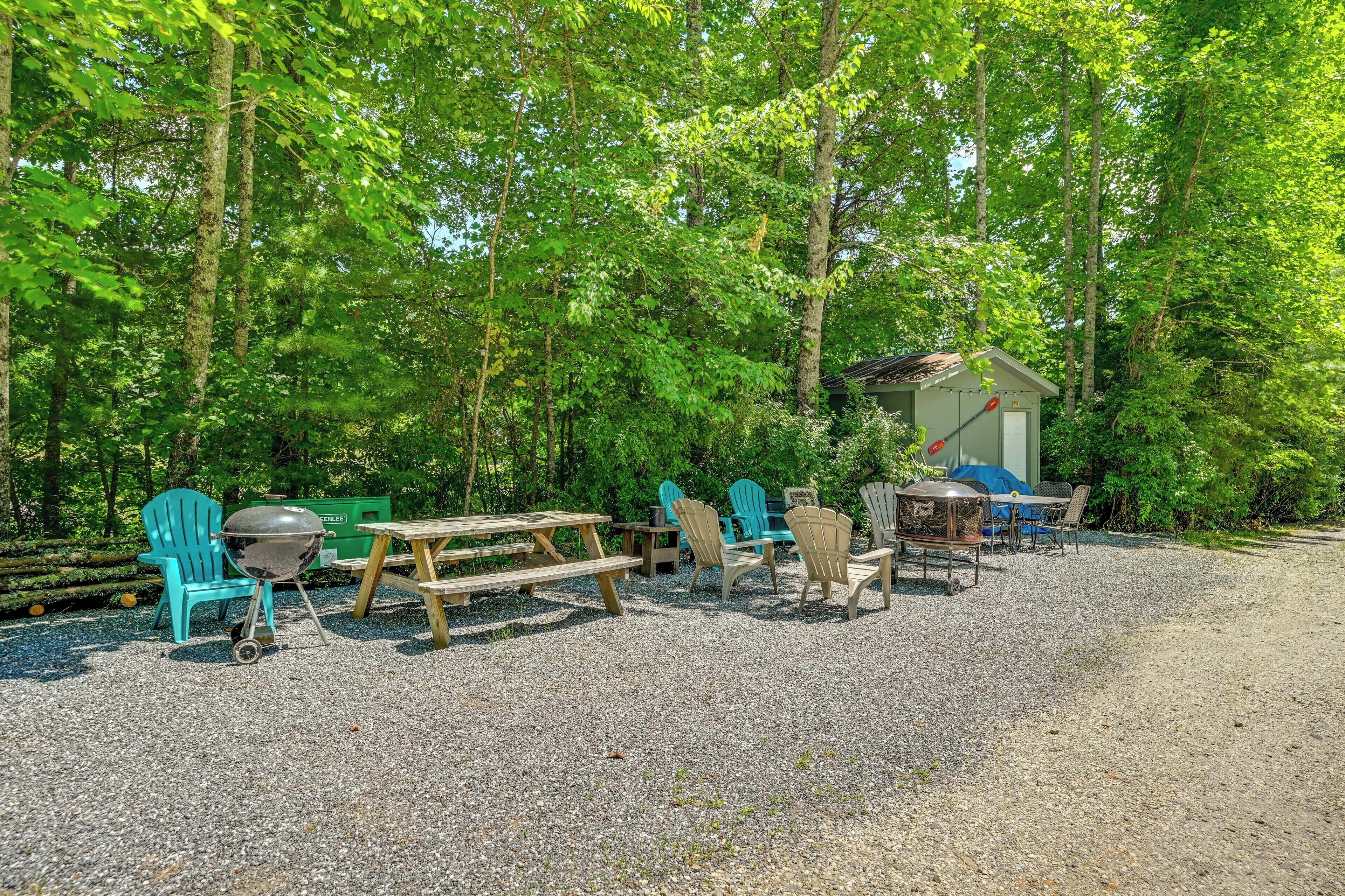 A picnic area with Adirondack chairs, a grill, and a small shed in a wooded setting.
