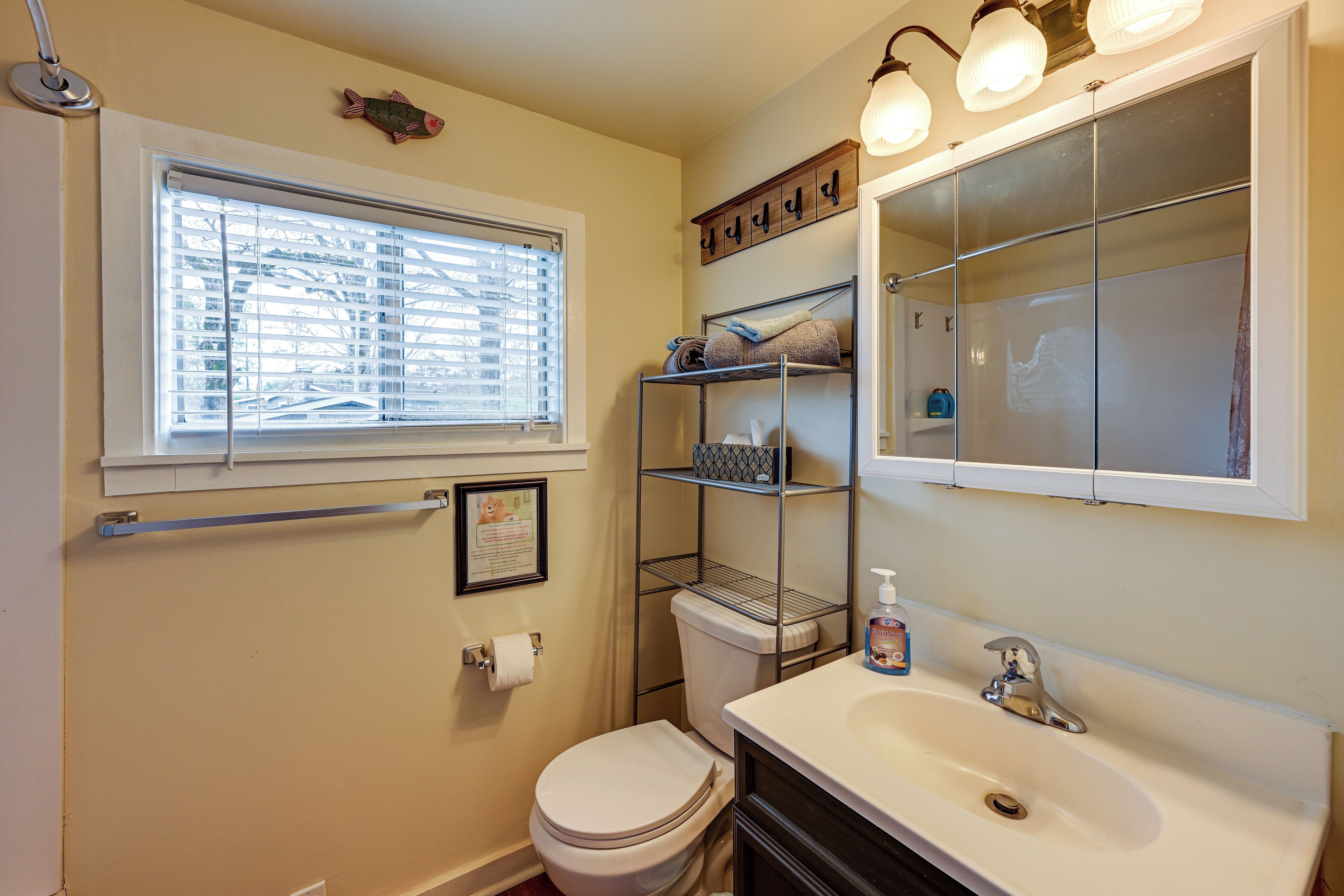 A bathroom with a toilet, sink, and window. There is a shelf above the toilet and a towel rack above the sink.