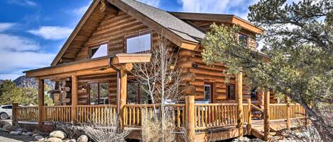 A log cabin with a porch, a car parked in front, and a mountainous backdrop.