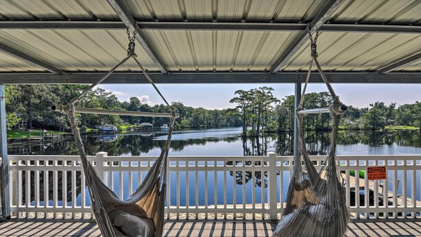 A hammock under a covered deck overlooking a lake with a white fence and a warning sign. - 'the Grey Goose': Waterfront Home With Beach (New Bern)